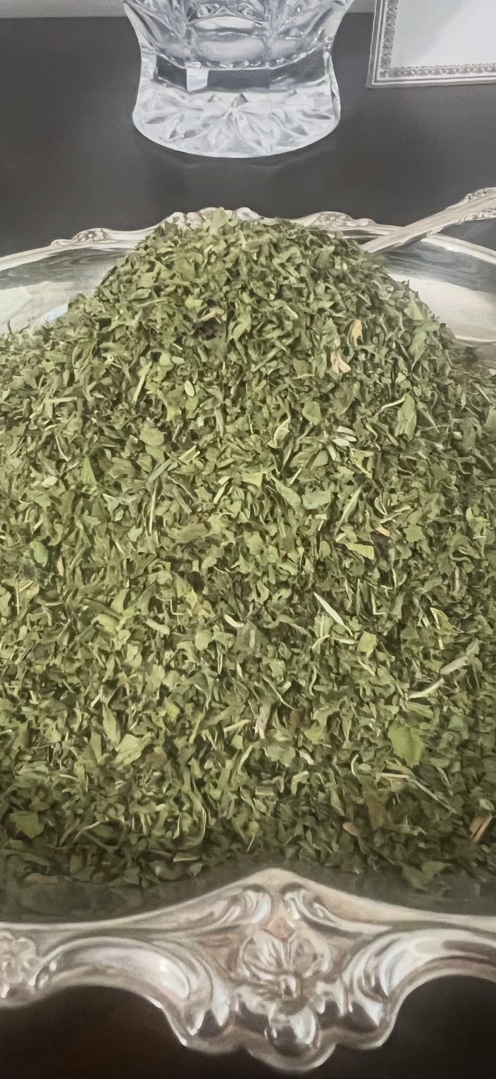 A pile of dried green leafy herbs or tea leaves displayed on an ornate silver-colored metal tray, with a small decorative spoon resting inside.